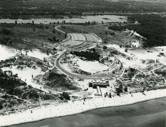 Historic black and white aerial photograph of West Beach as viewed from above the lake with the bath house in the lower center. Sandy dunes and woodland surround.