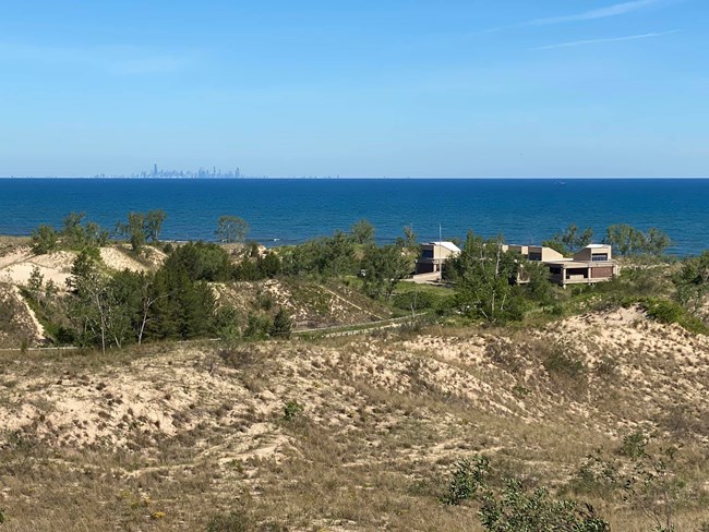 Elevated view of West Beach's coastal dunes along the lakefront. A concrete building, the West Beach Bathhouse, stands near the shore. The Chicago skyline is visible on the lake beneath a beautiful blue sky.