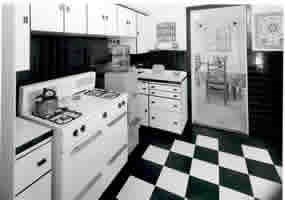 black & white photo of kichen with stove, water cooler, black and white checked tile floor and cabinets.