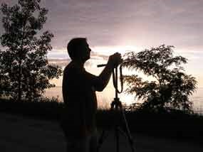 silhouette of man standing behind a tripod with camera on to, trees and sky in background