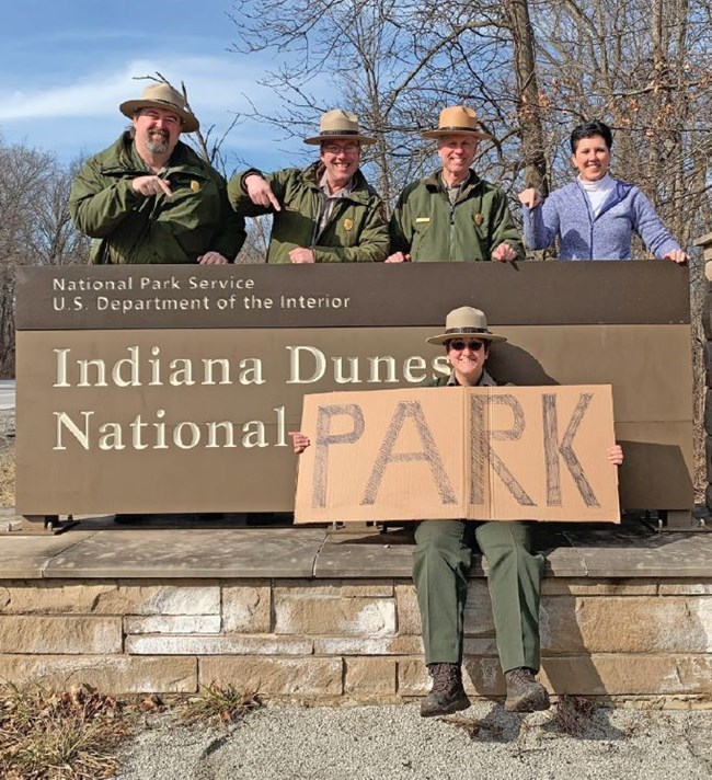 Group photo holding cardboard park sign over Indiana Dunes National Park road sign.