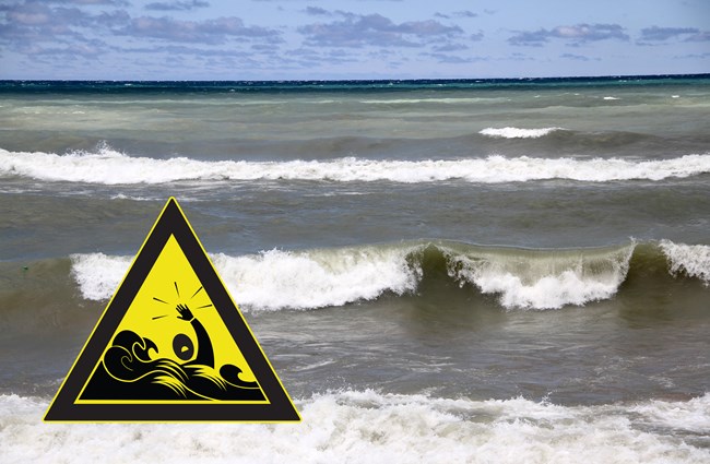 Gray waves with white caps crash at the nearshore of Lake Michigan. A yellow triangular warning icon is superimposed on the bottom left. The warning depicts a swimmer struggling in high waves.