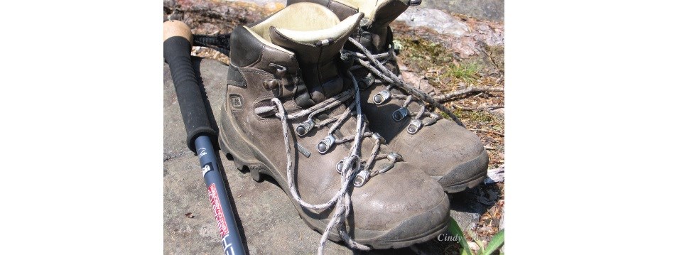 A photograph shows two well used brown hiking boots on a rock next to the handle of a hiking pole