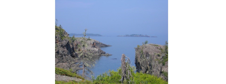 A calm cove is surrounded by angular rock, with rocky islands in the distance