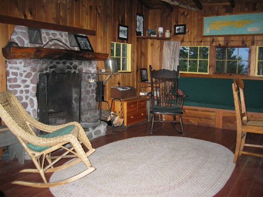 A photograph shows a rustic cabin with a large stone fireplace, chairs, and a couch surrounding a large circular rug