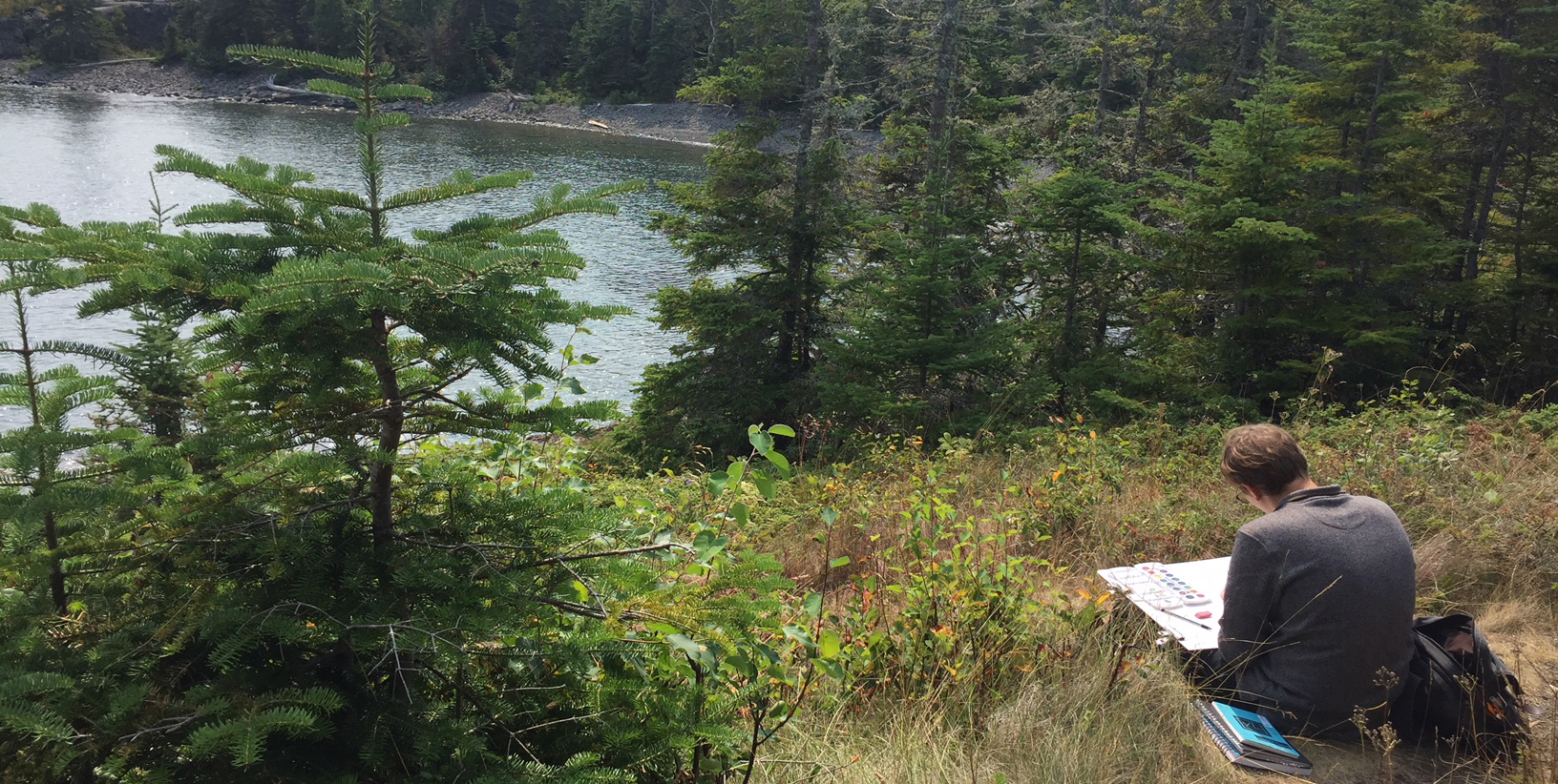 An artist works on a painting while sitting on a trail near a lake.