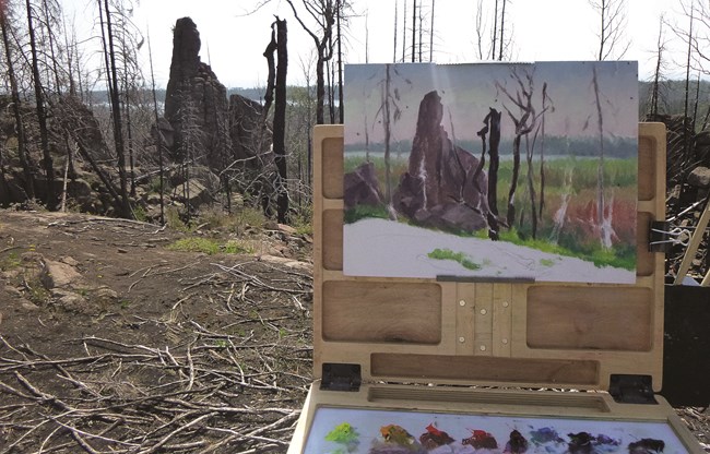 An easel with a painting in progress on it is set up in a burned landscape. The subject of the painting is in the background.