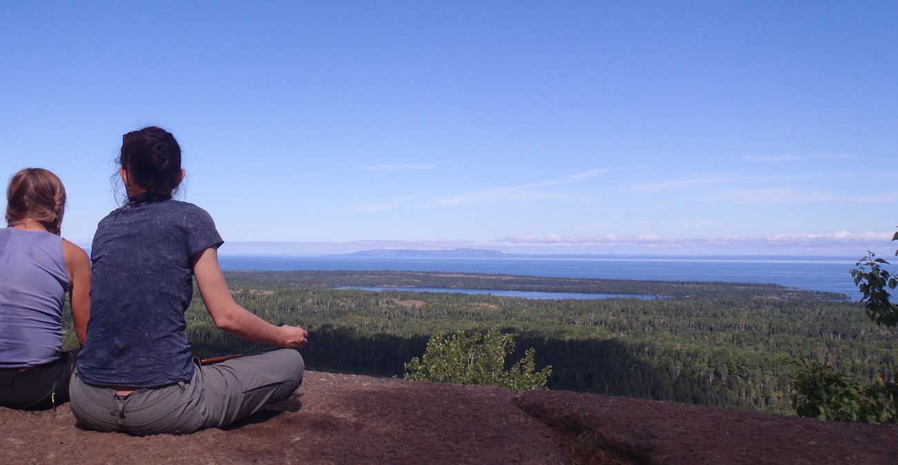 Two people sit on a rock and view a scenic landscape from above.