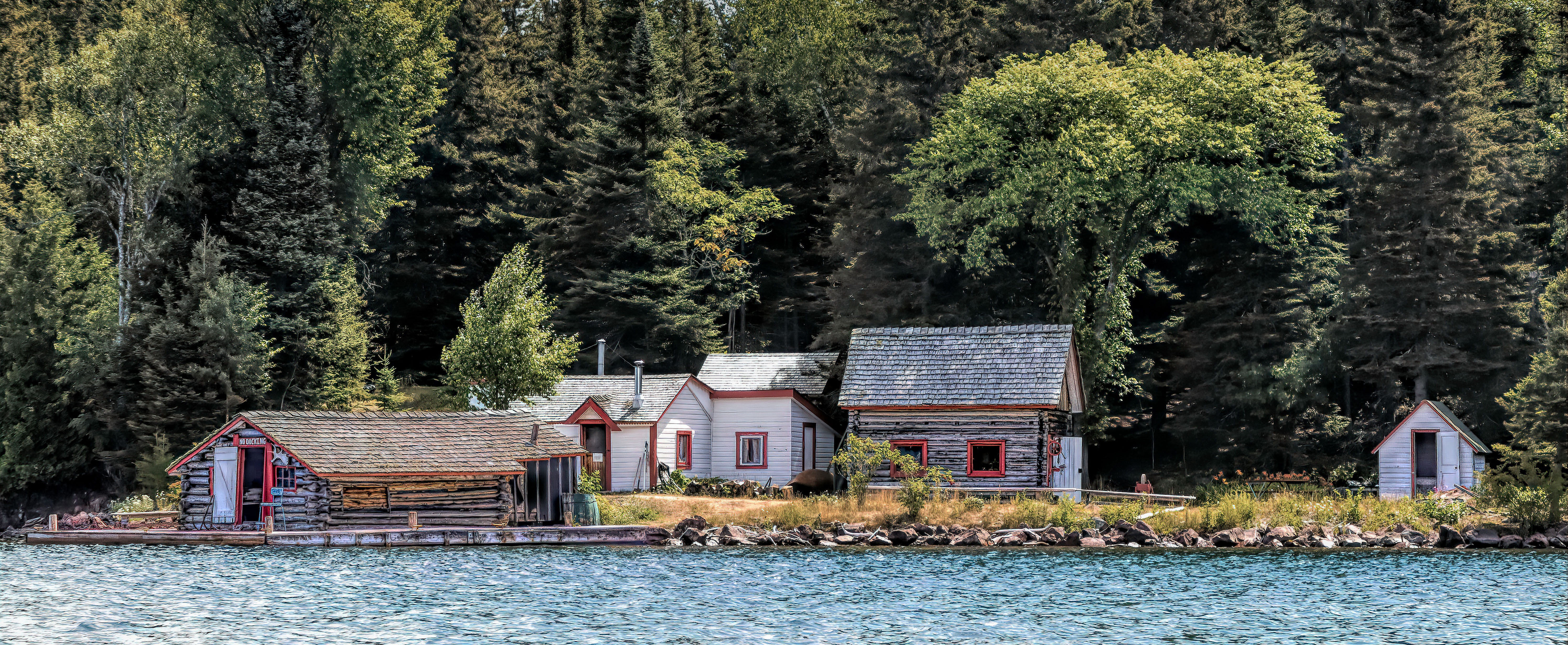Multiple grey historic cabins and a lakeshore surrounded by forest.