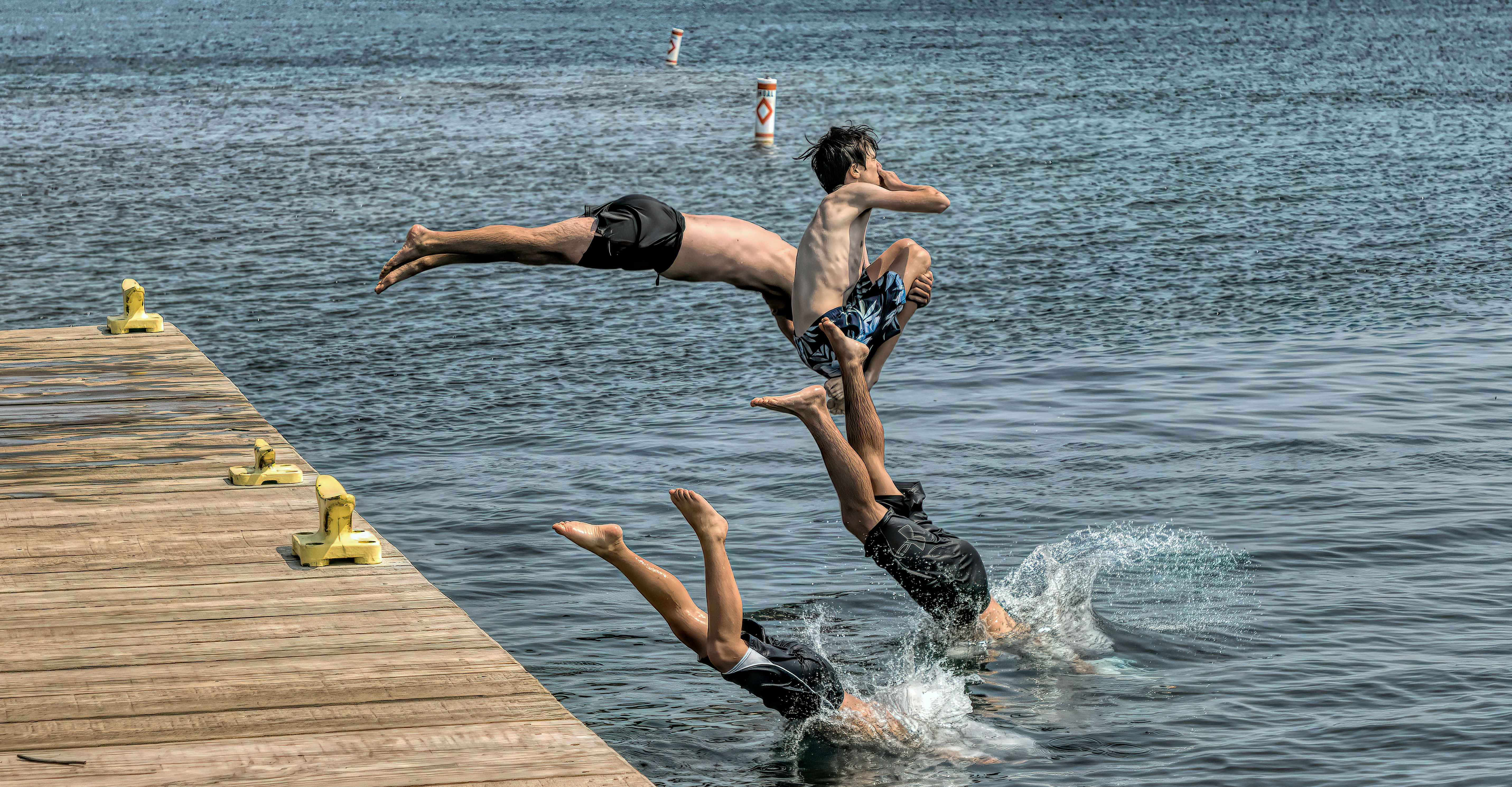 Three people jump and dive off a wooden dock into a lake.