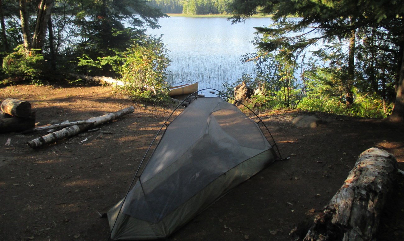 A small tent pitched at a campsite on a lake with a canoe at the shore.