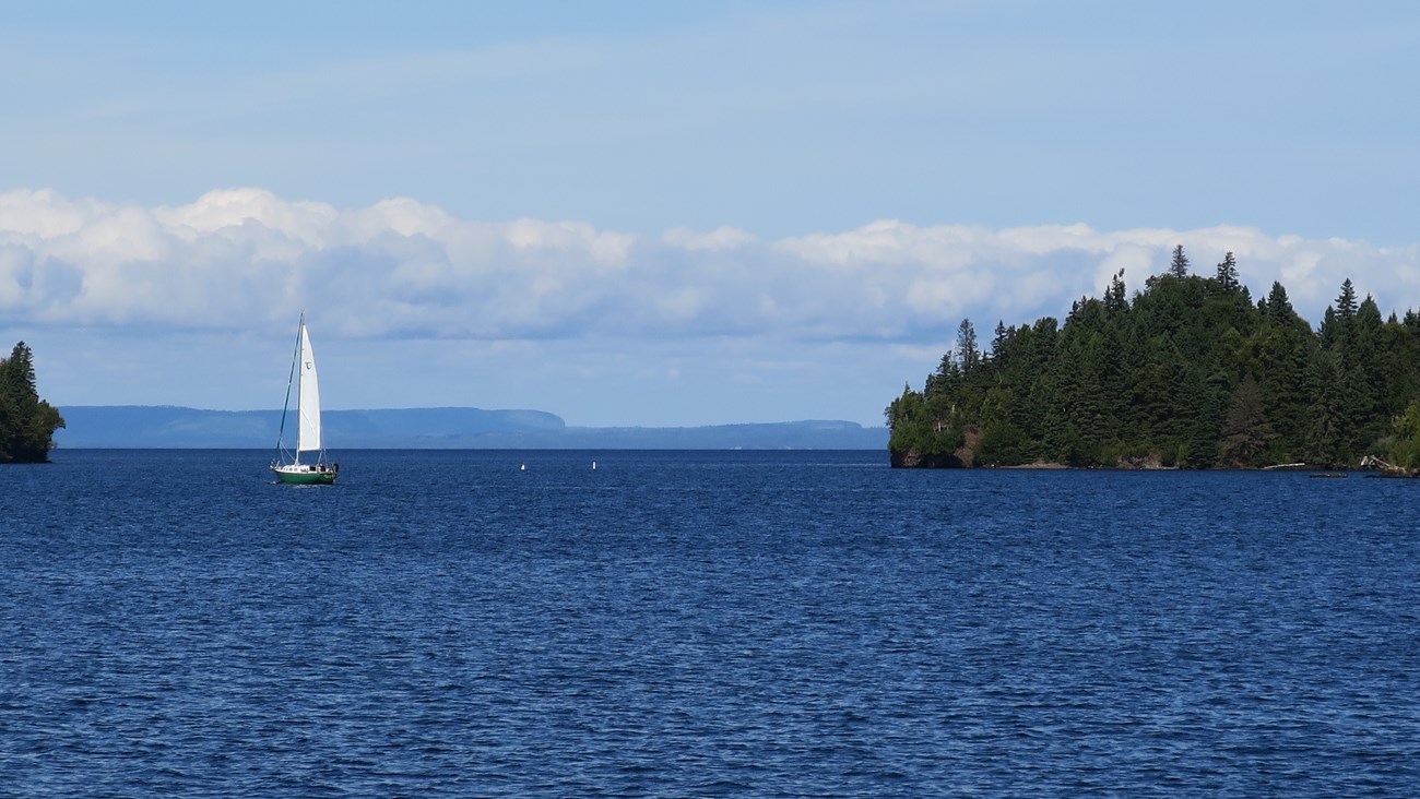 A sailboat in the water on a clear day, sailing towards a small island.