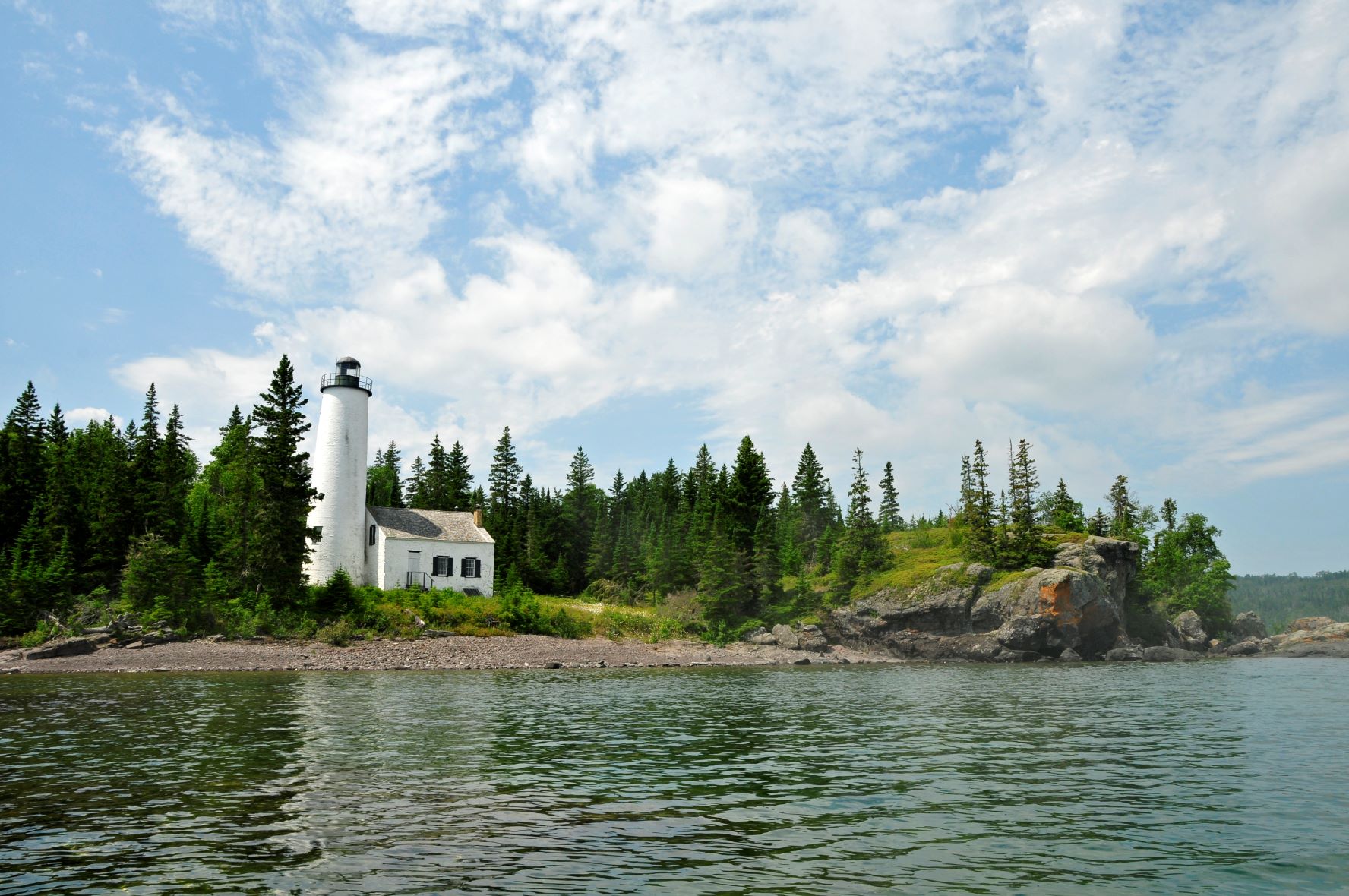 Rock Harbor Lighthouse on a rocky shoreline surrounded by boreal foreal. Puffy clouds speckle the blue sky. Calm Lake Superior ripples in the foreground.
