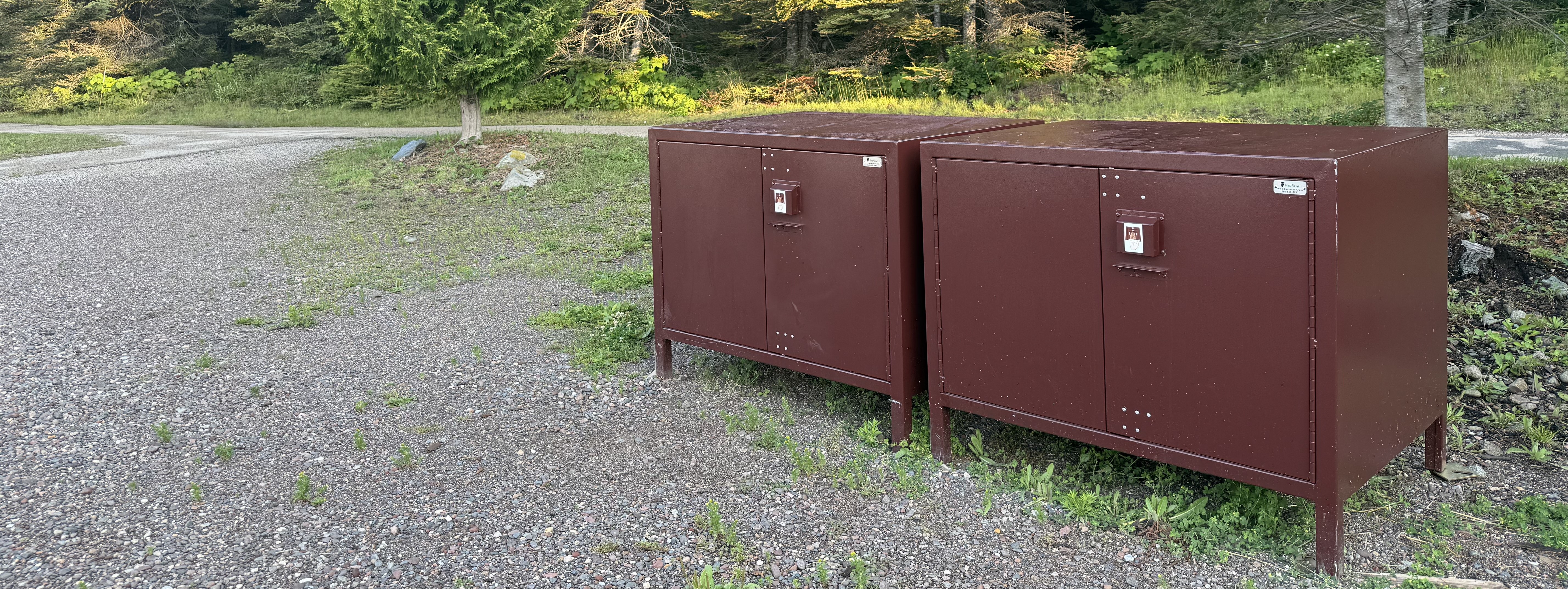 Two large, brown, metal lockers on a gravel path near a forest.