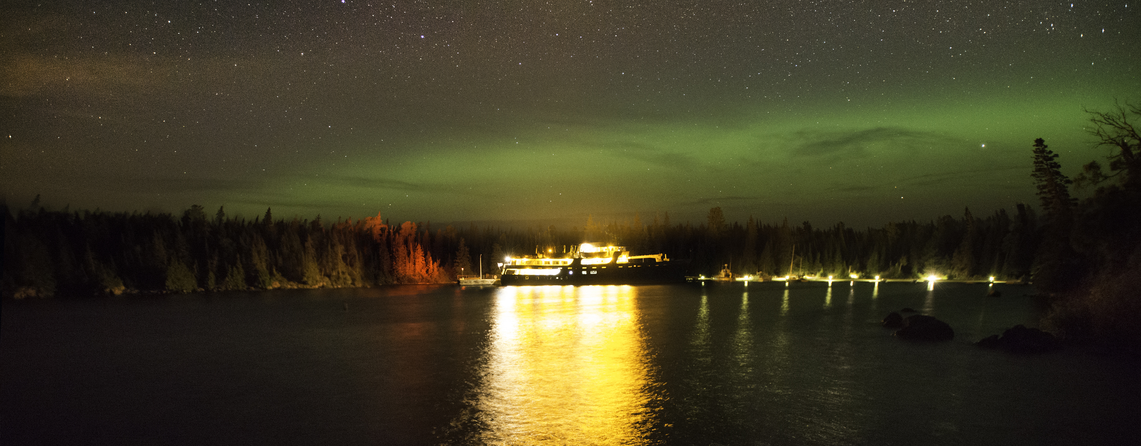 Ranger III Ferry at dock at night in Rock Harbor. Lights reflect of the of the dark water. Green waves of the aurora borealis dance above the treeline against a star-filled sky.