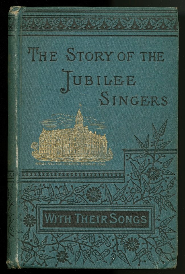 A hardcover book titled “The Story of the Jubilee Singers: With Their Songs” written by J. B. T. Marsh. The cover is blue with black text. A black floral pattern is on the bottom half of the cover. A gold depiction of Jubilee Hall at Fisk University is at