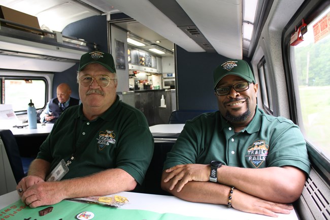Two men wearing green NPS Volunteer polos and ball caps sit on an amtrak train, smiling at the camera