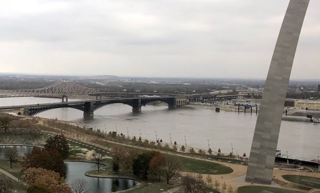 A brownish river, with a black arched bridge crossing it. Green plants in the foreground. A sloping leg of a gigantic steel arch is on the right side.