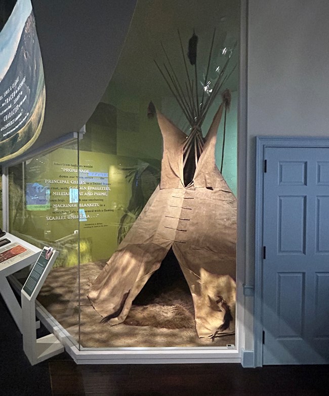 A cone-shaped tipi behind glass in a museum. A blue access door is to the right.