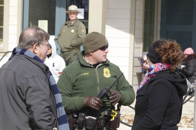 Outside during the day. Law enforcement ranger stands with a radio in hand. Woman in black jacket on right. Man in gray coat on left.