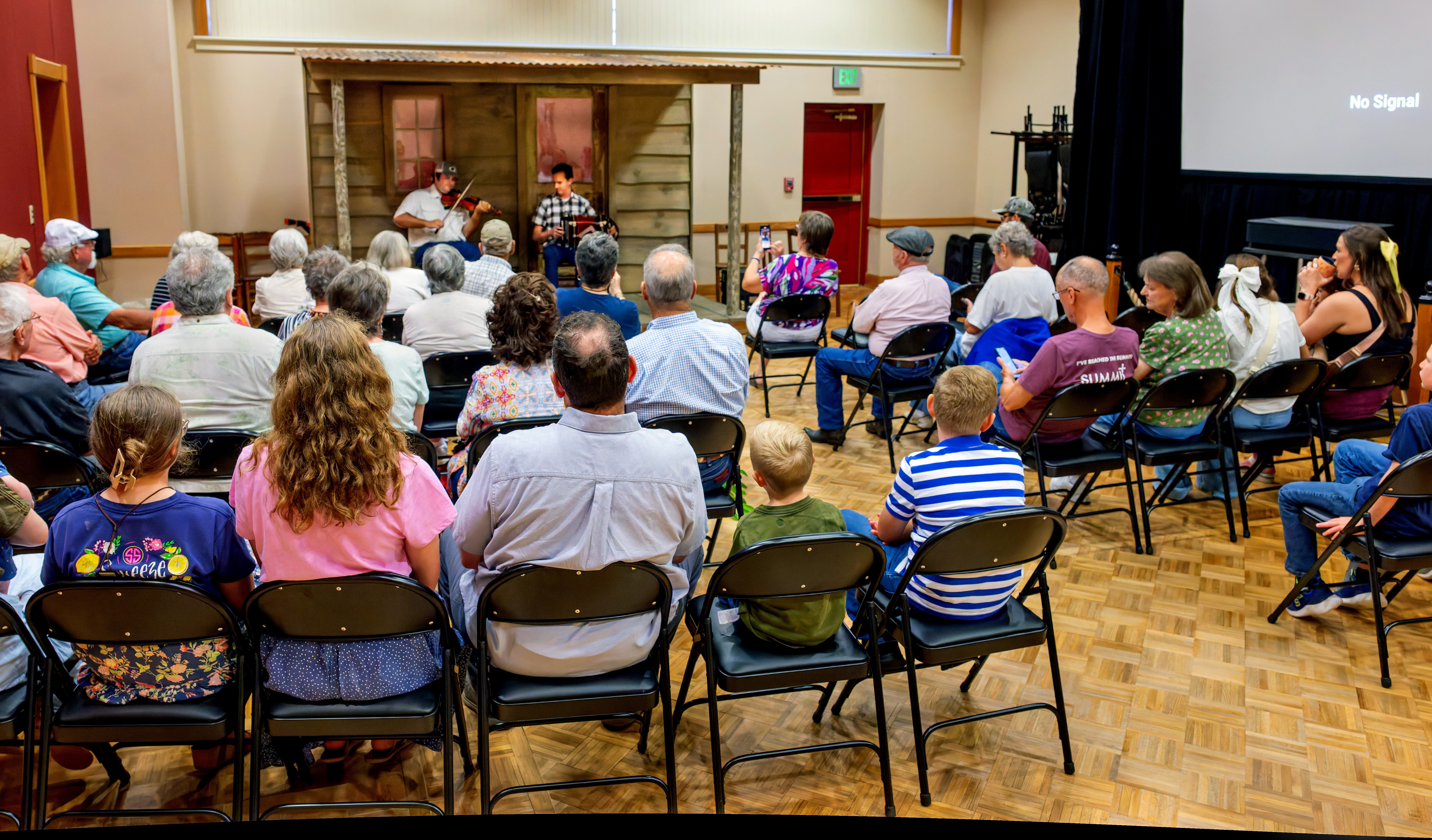 An audience of people sitting and listening to two musicians perform on a front porch stage.