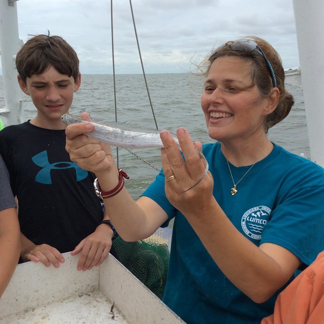 A young woman smiles, holding a small thin silver fish up while a young boy smiles watching her work.