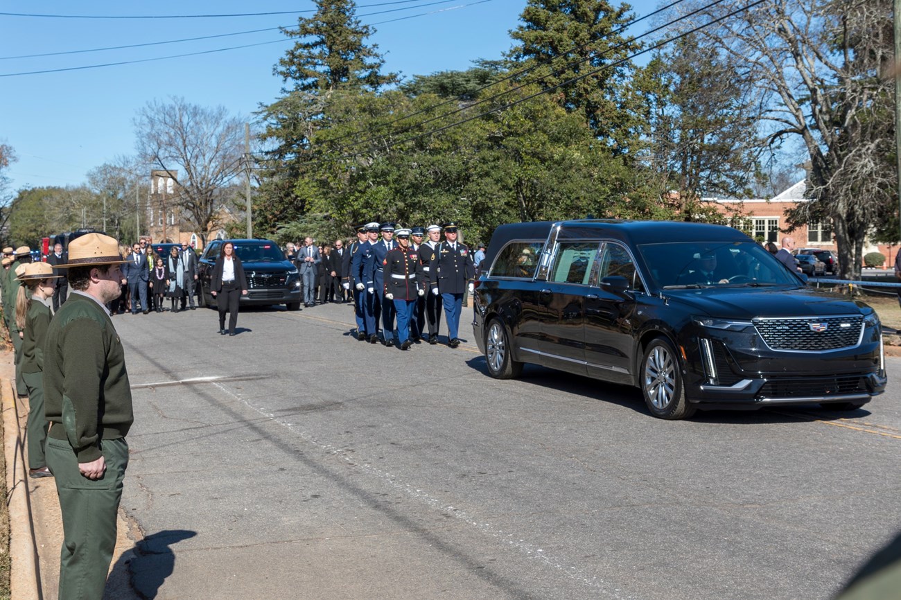 Hearse traveling down a street lined with park rangers