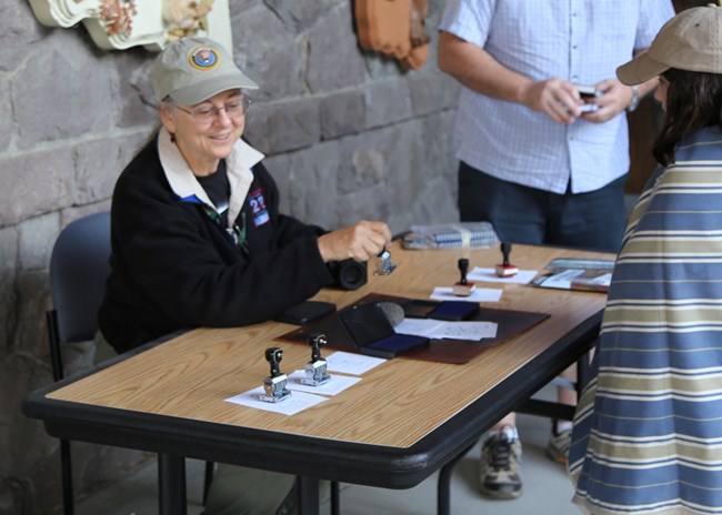 A girl watches a woman stamp pieces of paper at a table.