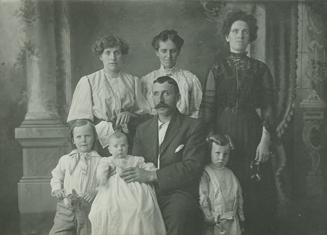 A black and white photo of a man in a suit sitting and holding a baby, two children next to him, and three women standing behind.