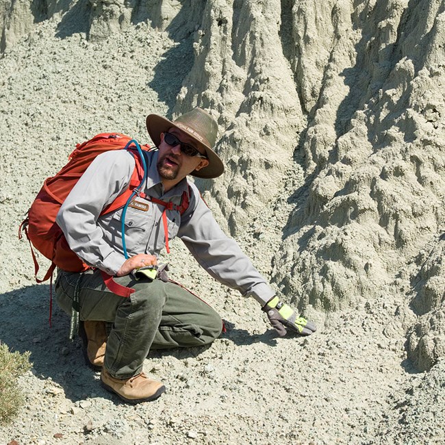 A man in a Park Service uniform, wearing a red backback, crouches in front of a blue-ish, grey-ish green rock exposure. He balancing on one hand to the right of the frame.