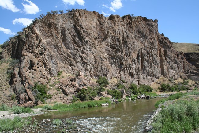 A dark brown and black outcrop of rock rises above a river.