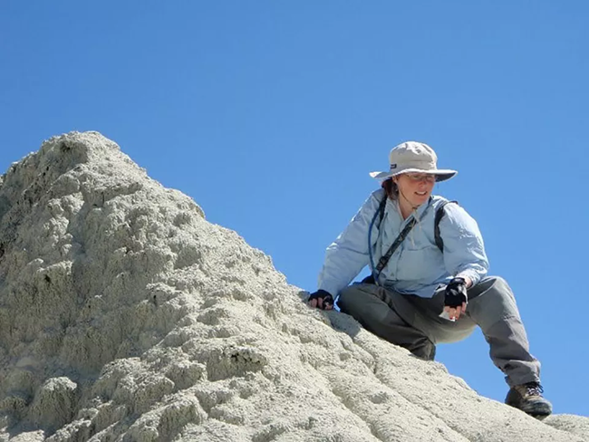 A woman in long sleeves, pants, hiking boots, and wide brimmed hat crouches on a rock.