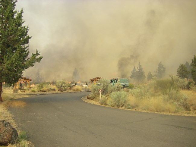 Thick black smoke rising from vegetation beyond a parking lot. A faint flicker of flames can be seen. Fire fighters stand nearby.