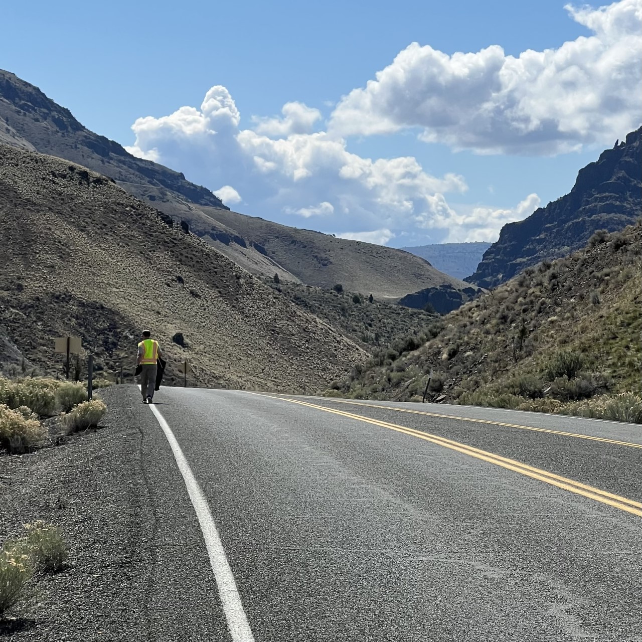 A person wearing a reflective vest walking along a highway shoulder.