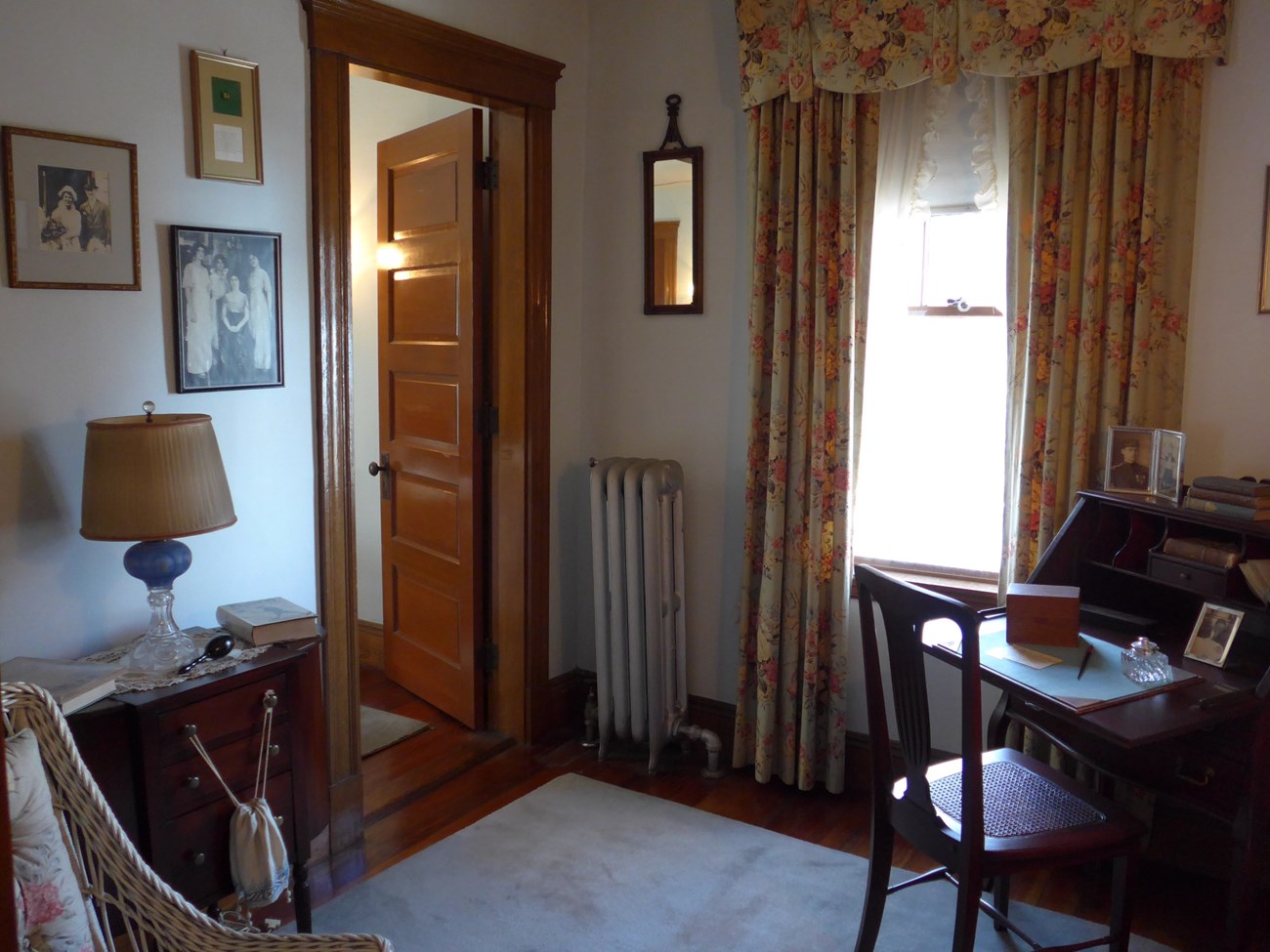 A photo of a small room. A wooden desk and chair sit by a curtained window. On the opposite side, a table holding a lamp, books, and a darning bulb stands by an open door. Black and white family photos hang on the wall.