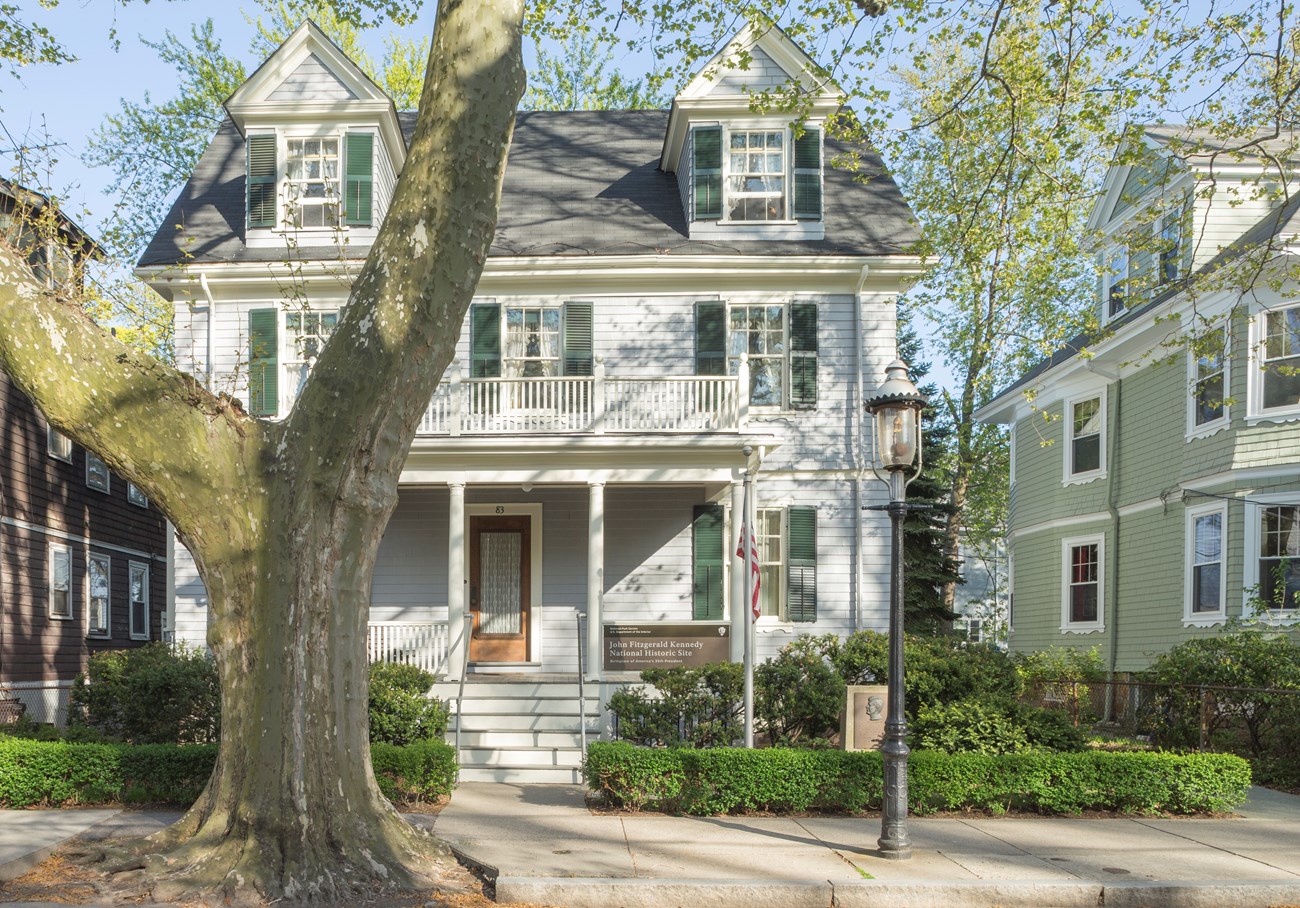 The front of a grey, two story house, partially obscured by a tree, viewed from the street.