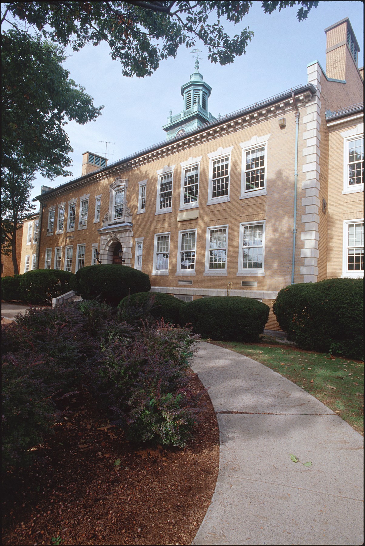 A photo of a wide, two story building made of tan brick, with twenty seven visible windows, a light blue bell tower with a clock on it, a front door set in an archway with steps, and manicured round hedges and a walkway in front.