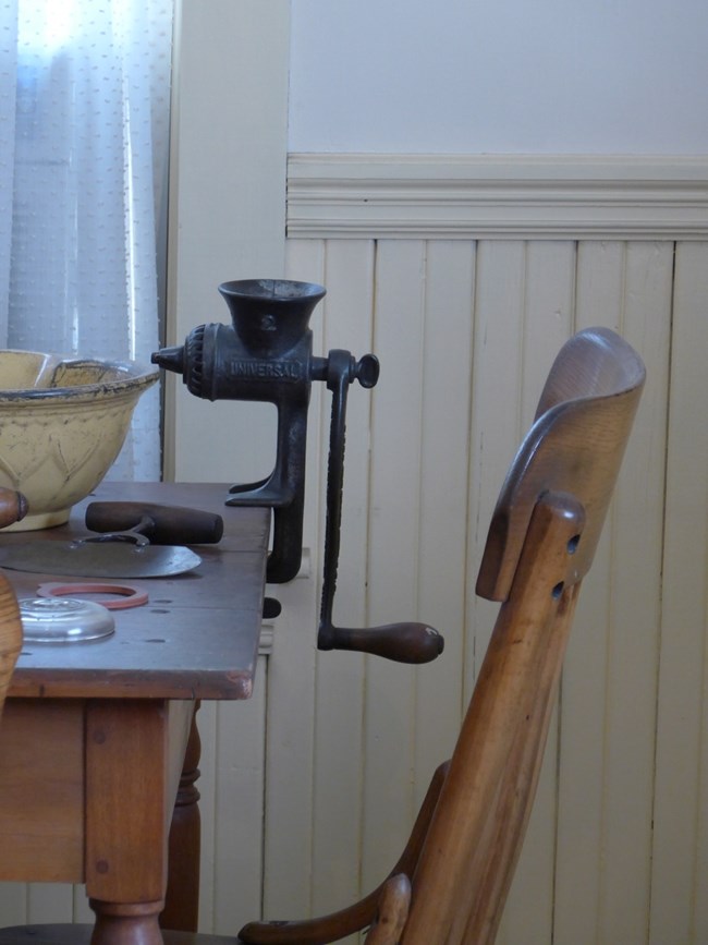 A photo of a metal food chopper clamped to a table, consisting of a funnel, grinding chamber, clamp, and crank. An axle runs through the chamber connecting the blade to the crank, which hangs parallel to the body and has a wooden handle.