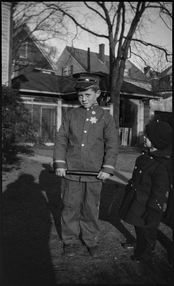 A black and white photo of a boy in a police costume holding a billy club, while a small girl in a coat looks on. They are standing on a lawn in front of a garage. Bare trees stand in the background.