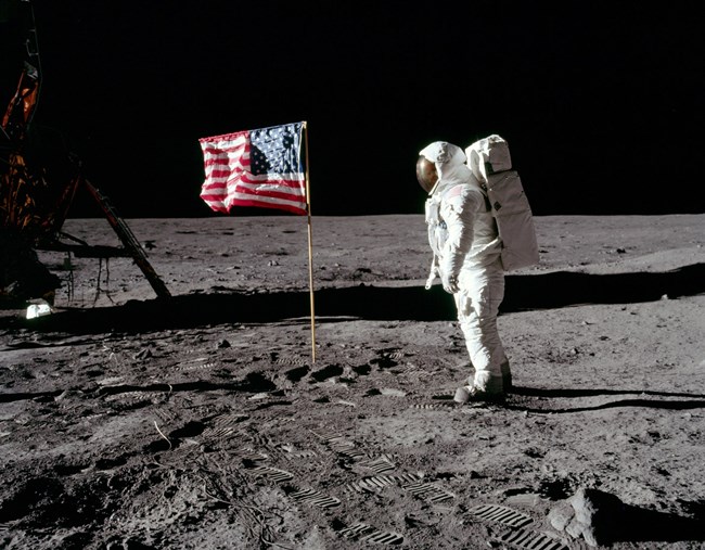 Astronaut on the right stands on the moon observing an American flag on the left