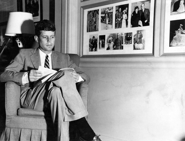 a black and white photo of JFK seated and reading.  Photos are on the wall to the right of him