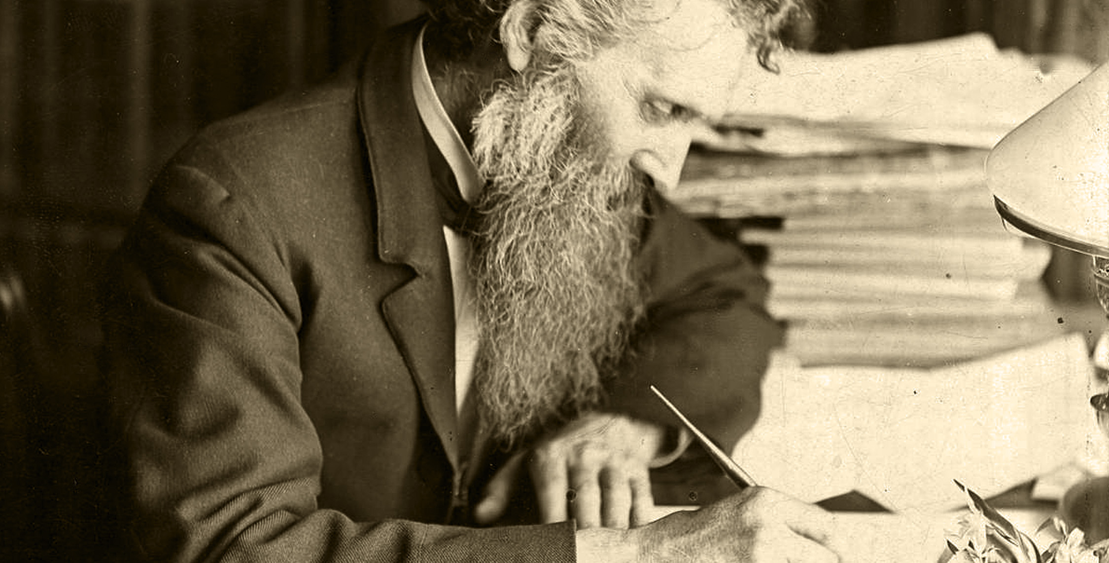 Historic photograph of John Muir seated at a desk, writing with a pen in hand. Papers and a lamp are visible on the desk, reflecting his dedication to documenting his thoughts and advocating for conservation.