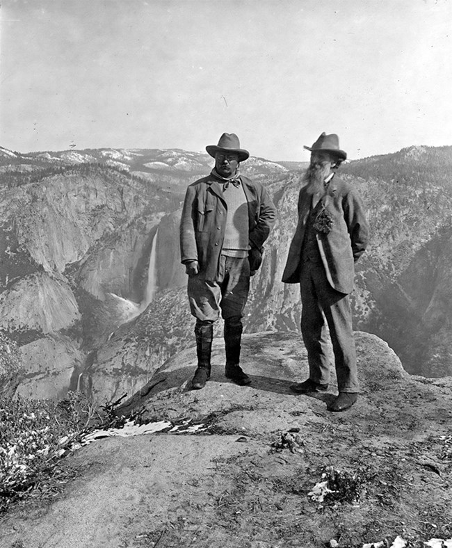 Black-and-white photograph of President Theodore Roosevelt and John Muir standing together in Yosemite National Park. Behind them, a waterfall cascades down rugged cliffs.