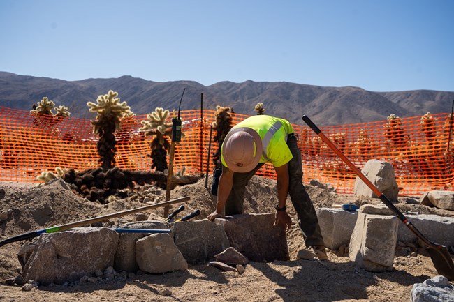 A trail crew member in a safety helmet lines large rocks next to the trail.