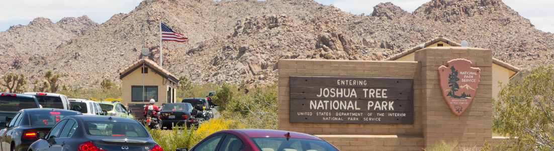 A line of cars waiting to enter the entrance booth passes a large brick sign that reads "Entering Joshua Tree National Park."