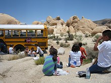 elementary school students sit outside in the park to eat lunch, with their school bus nearby