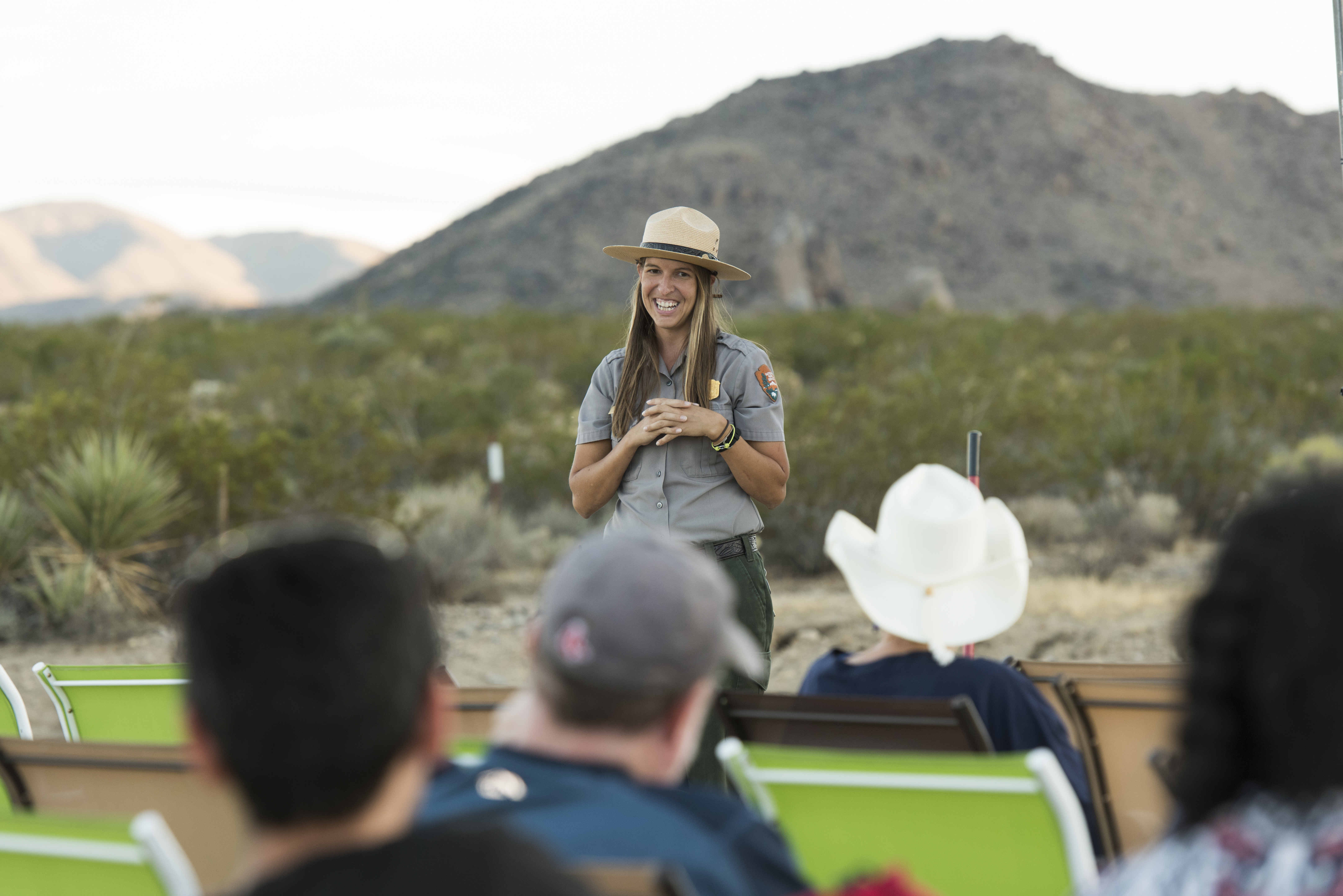 A ranger smiles in front of a crowd of seated visitors at sunset. Desert plants and rolling mountains fill the background.