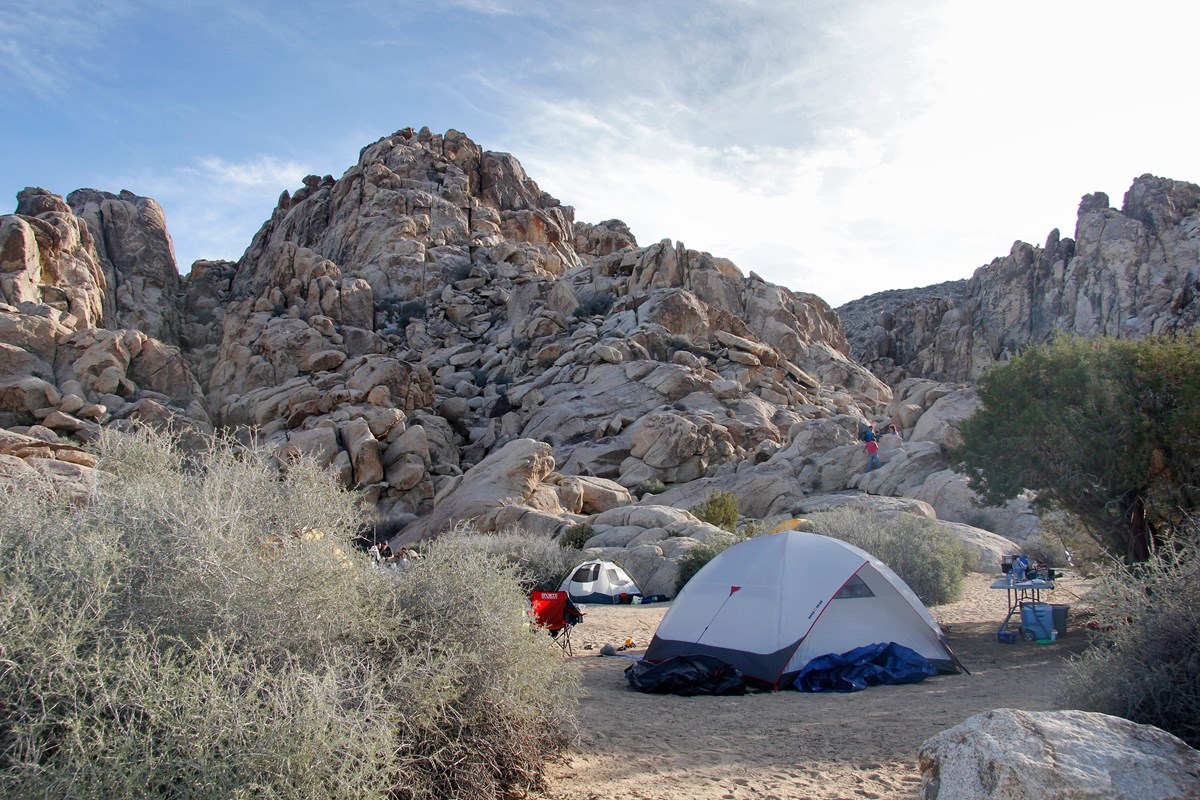 Color photo of a group campsite at Sheep Pass campground. One dome tent is in the right foreground, with a smaller tent in the background against massive rock formations. People are seen climbing the rocks to the right background.