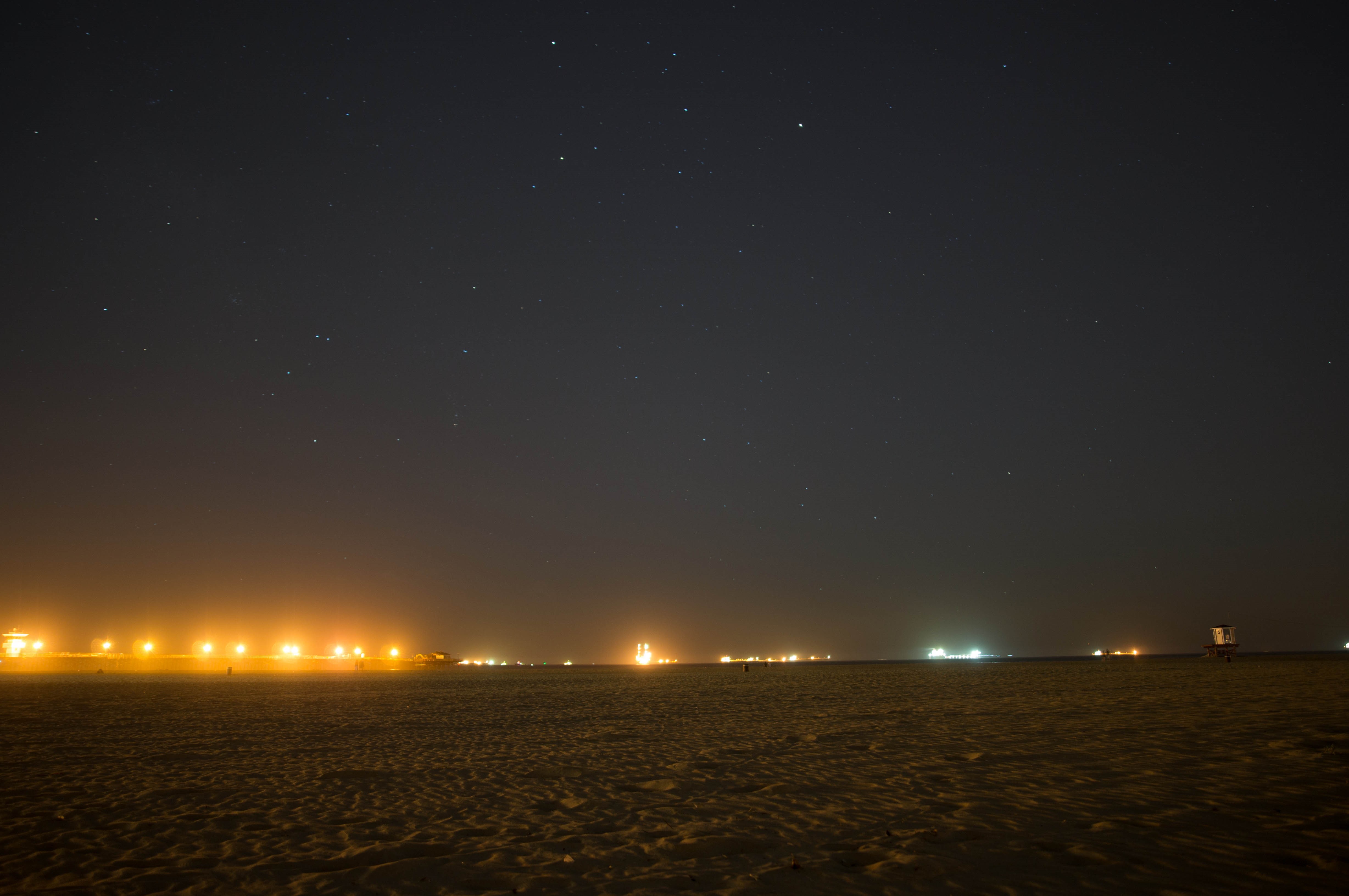 Faded stars shine over a brightly lit beach at night. The orange glow of the city is visible.