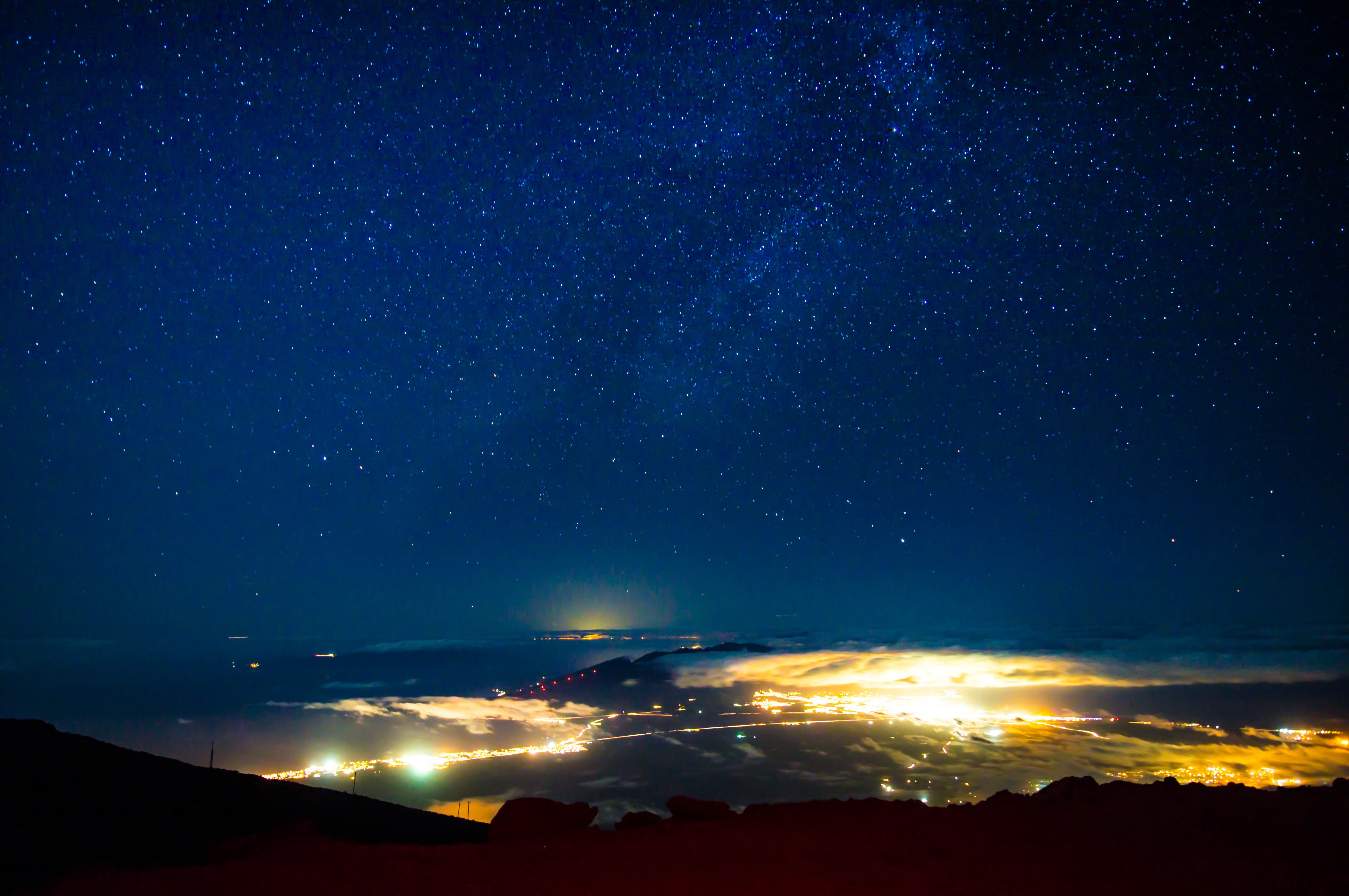 Bright stars in a cobalt blue night sky over the city of Maui. The city is lit up, but the light is contained by clouds. 